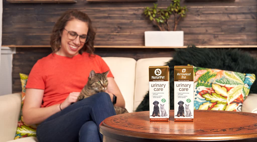 Woman sitting on a couch petting a tabby cat, with NaturPet Urinary Care and Arrowleaf Pet Urinary Care boxes displayed on a wooden coffee table in the foreground.