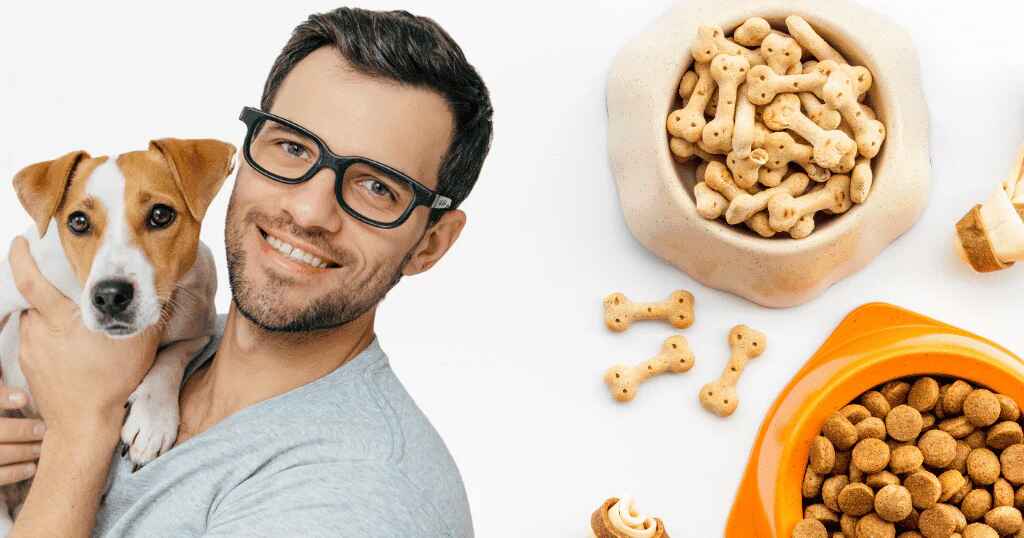 A man with glasses holding a Jack Russell Terrier with bowls of dog food and biscuits.